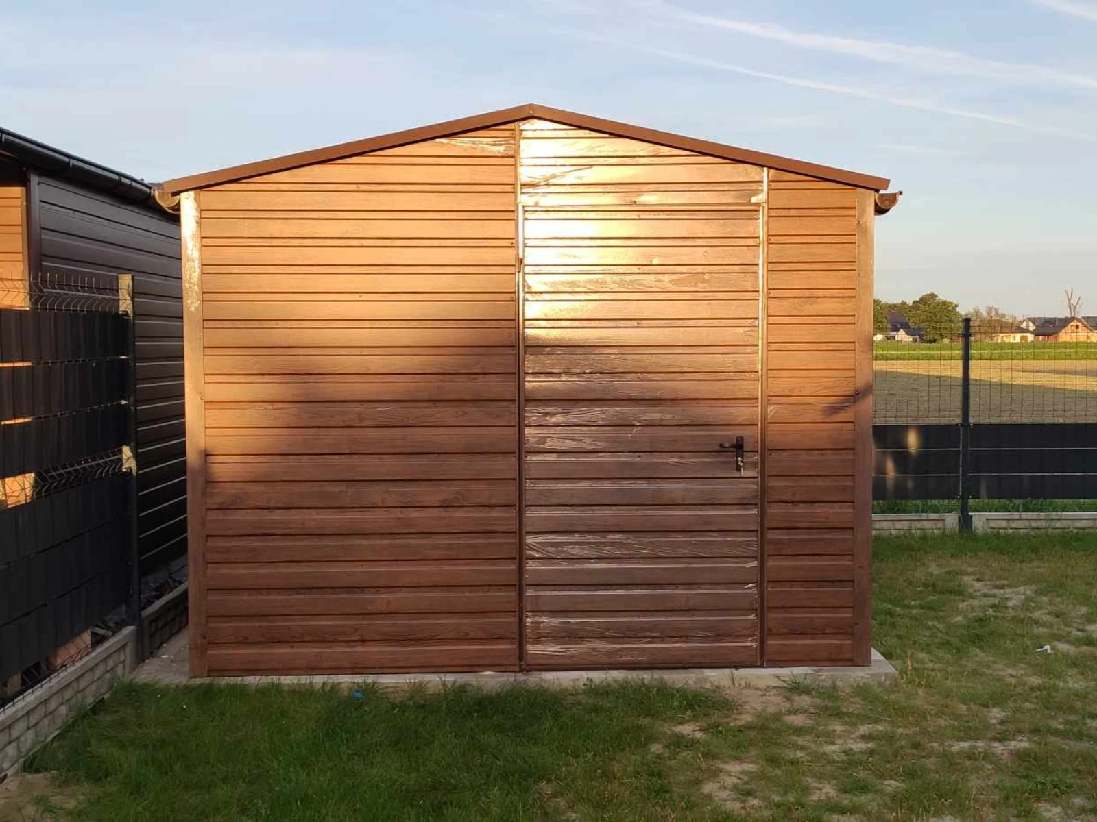 Domek narzędziowy z drewnopodobnych paneli stalowych na betonowej płycie w ogrodzie