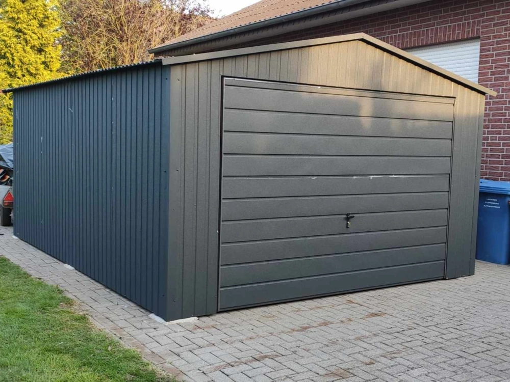 Szary stalowy garaż blaszany z dwuspadowym dachem i bramą segmentową na podjeździe