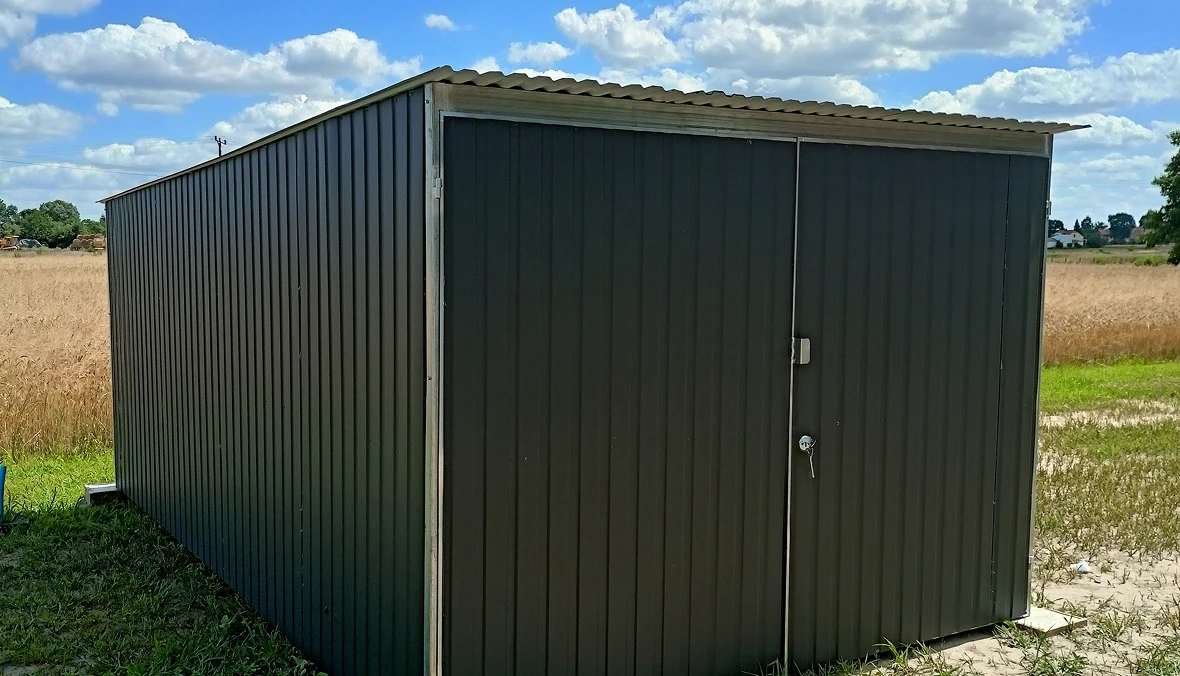 Czarny stalowy garaż blaszany z dwuskrzydłowymi drzwiami na tle pola