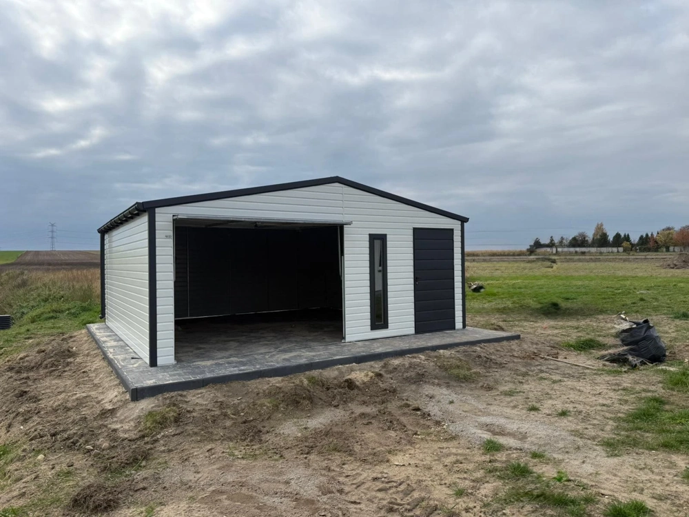Garaż blaszany z białym panelem i czarną ramą osadzony na betonowej płycie na otwartym terenie