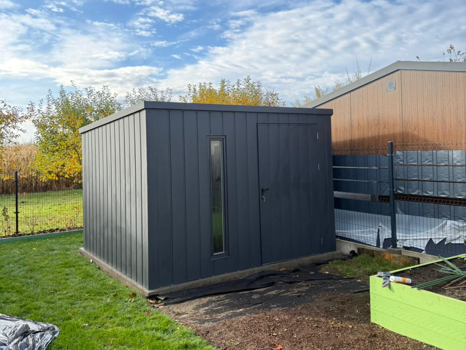 Stalowy, ciemnoszary domek narzędziowy z drzwiami i wąskim oknem na betonowej podstawie w ogrodzie