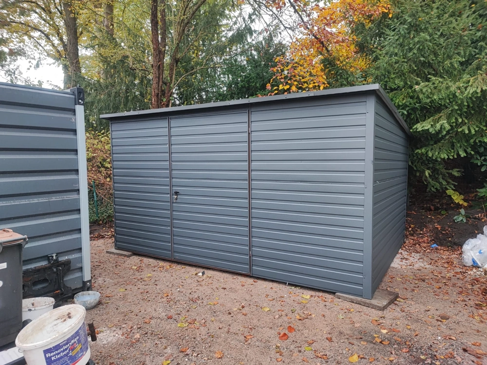 Szary garaż blaszany z dwuskrzydłowymi drzwiami na żwirowym podłożu, otoczony drzewami