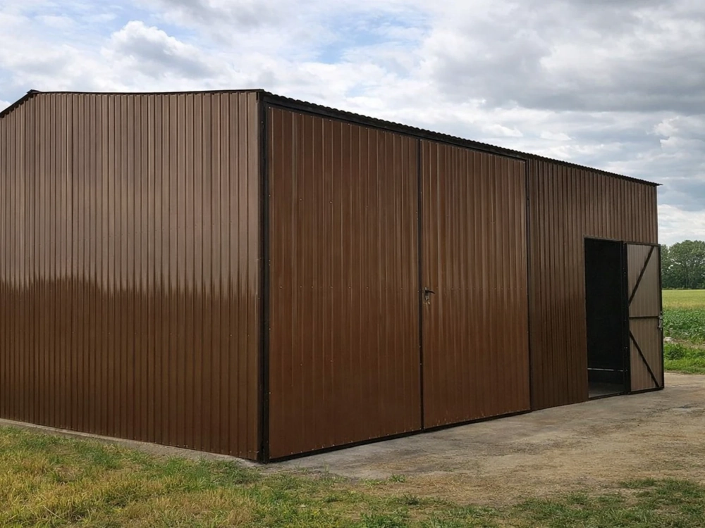 Brązowa hala blaszana z rozsuwanymi drzwiami, zamontowana na betonowej posadzce w otoczeniu zieleni