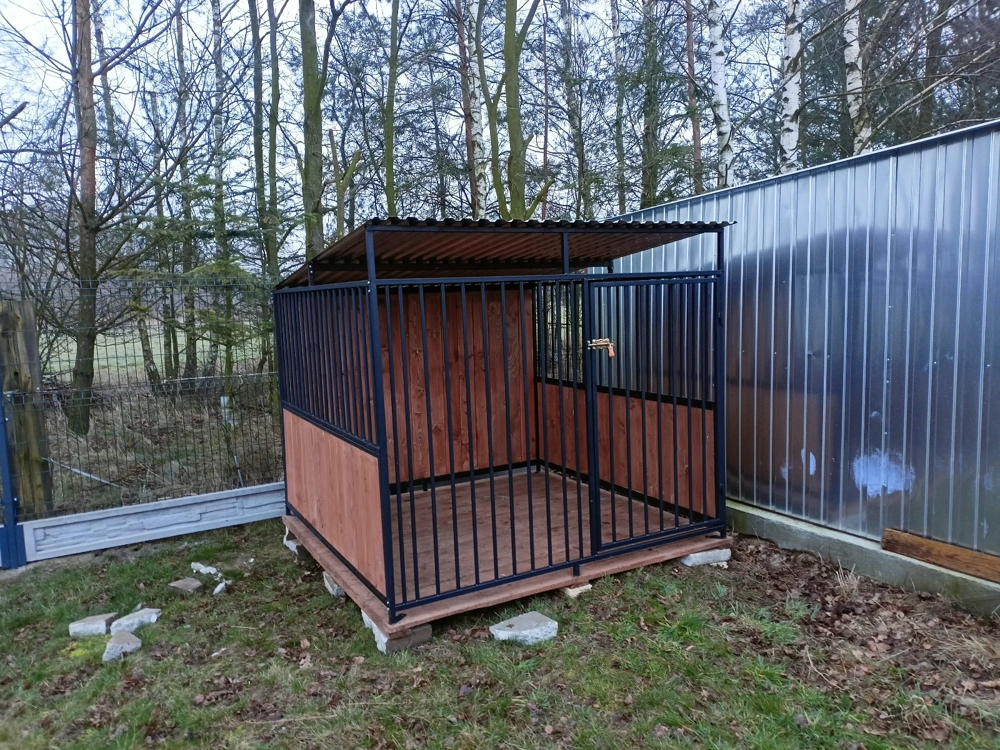Stalowa klatka kennelowa z drewnianymi panelami, podłogą i zadaszeniem, ustawiona na trawie