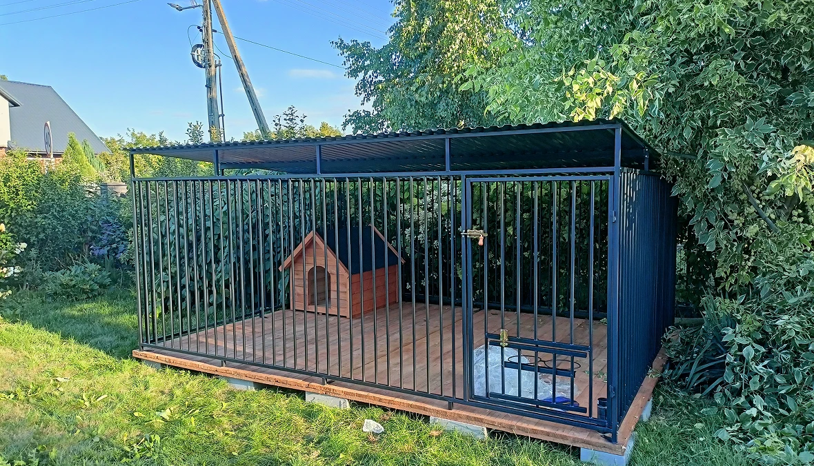 Metalowy, zadaszony kojec dla psów z drewnianą podłogą i budą, zamontowany na betonowych ławach, w ogrodzie